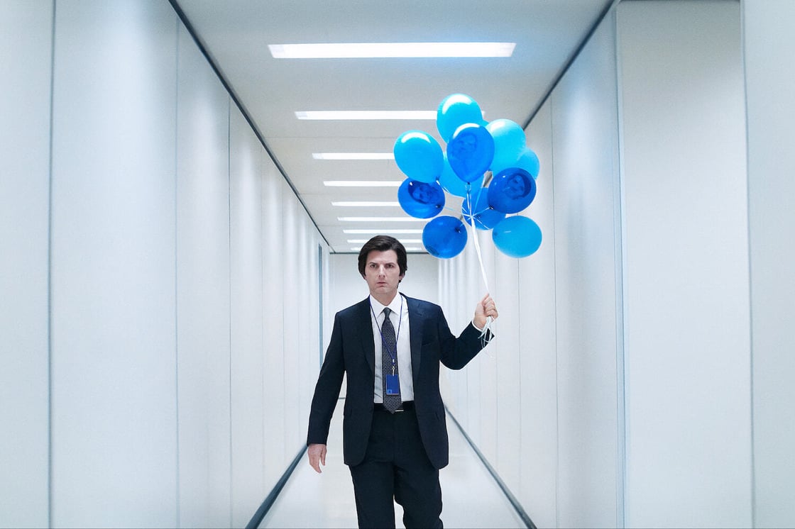 A screencap from Severance; Mark walks in a hallway with a bouquet of blue balloons