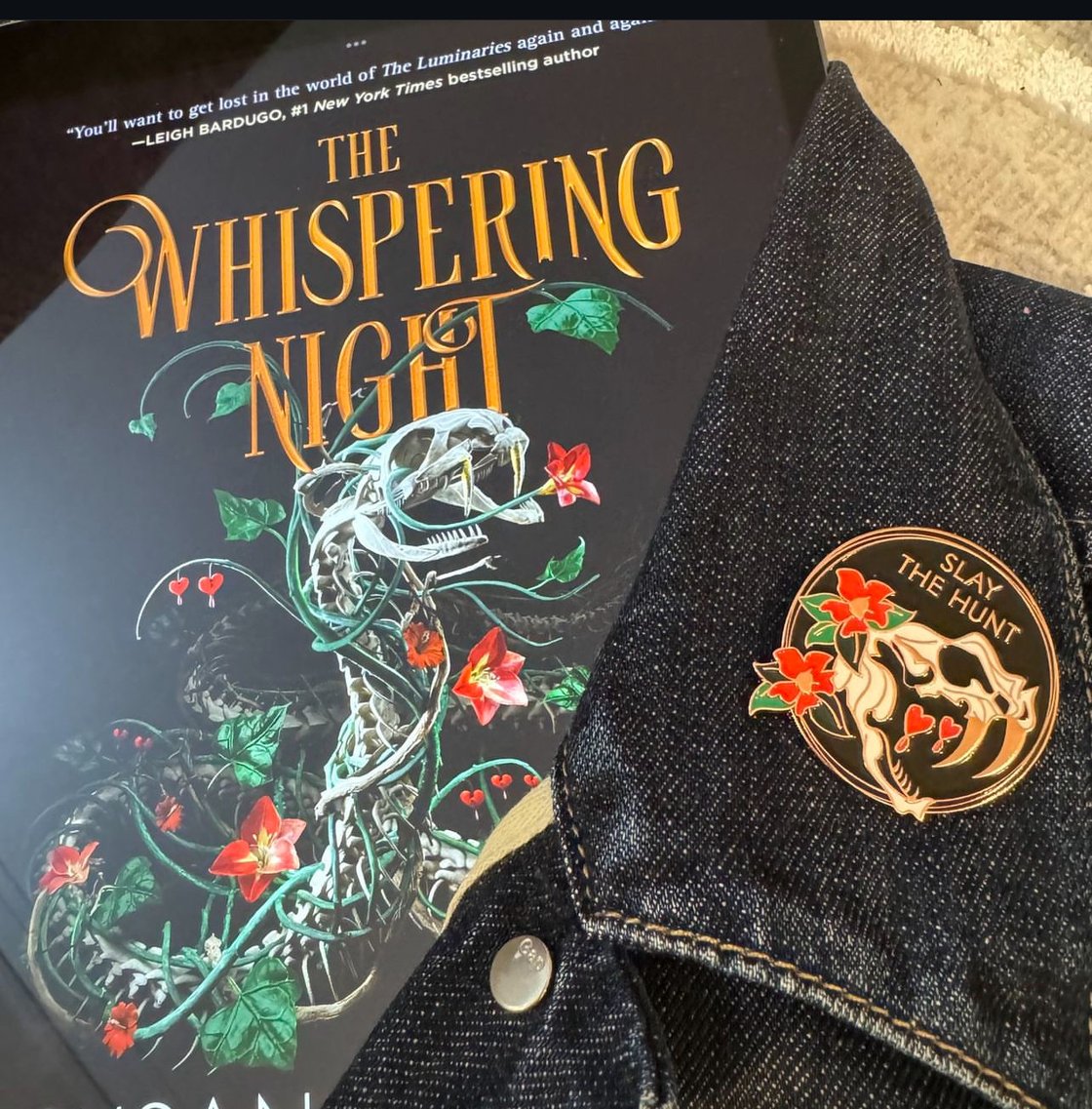 A photo of the hardcover edition of The Whispering Night by Susan Dennard—wrapped in a denim jacket featuring a "Slay the Hunt" enamel pin.