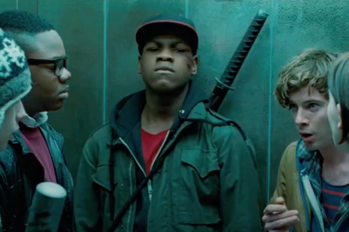 A screenshot from Attack the Block. The characters stand around talking against a wall, John Boyega staring past the camera with a katana on his back