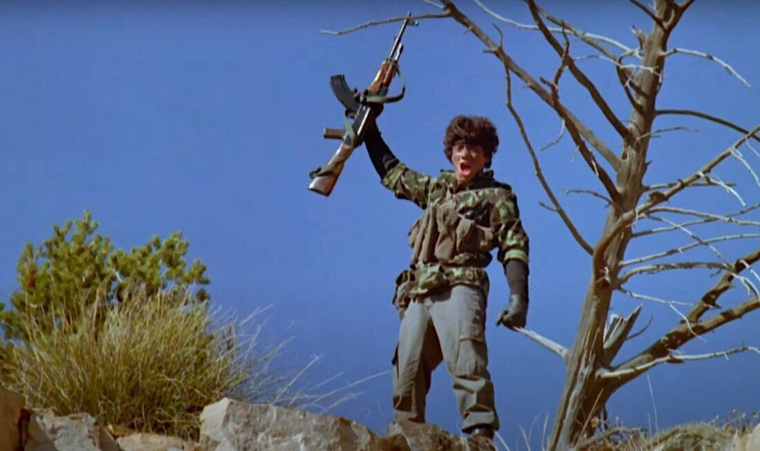 A screencap from Red Dawn. A man stands on a hill, yelling and holding a gun aloft