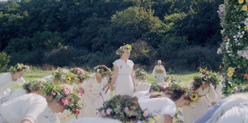 A scene from Midsommar. Florence Pugh stands in a white dress as a crowd of people bow around her